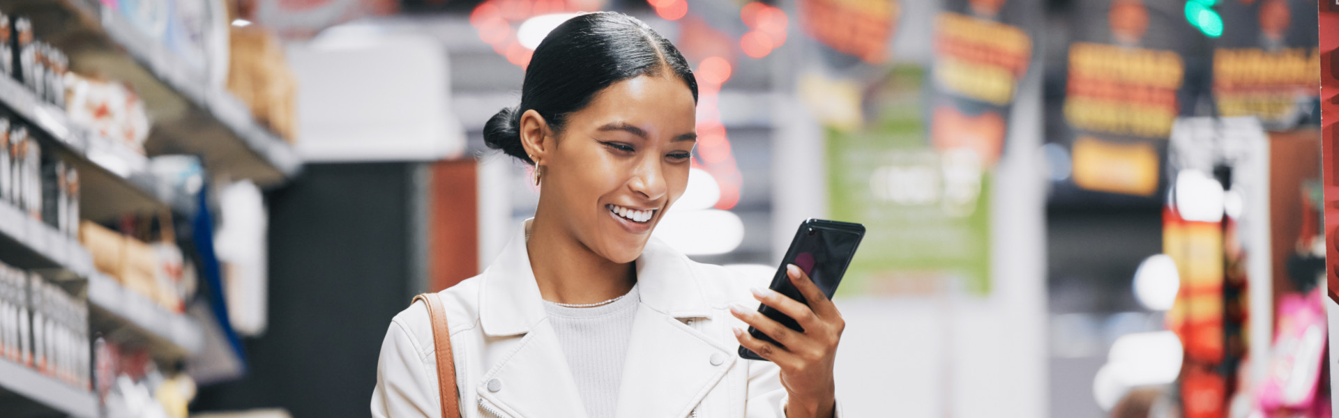 Junge Frau mit Einkaufswagen blickt lächelnd auf ihr Smartphone im Supermarktgang.