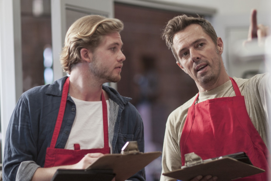 Zwei Supermarktmitarbeiter mit roten Schürzen und Klemmbrettern im Gespräch, einer zeigt nach vorn.<br />

